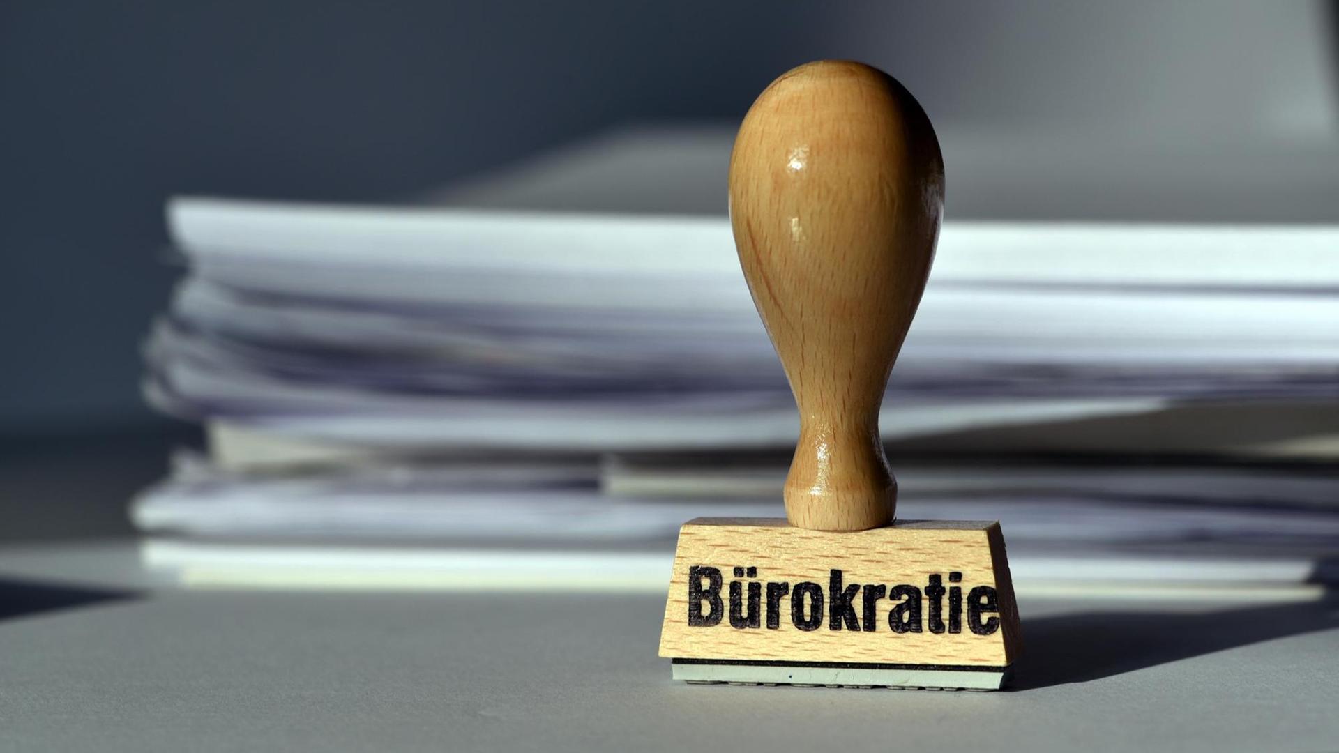 Ein Stempel mit der Aufschrift "Bürokratie" steht vor einem Stapel Papier.