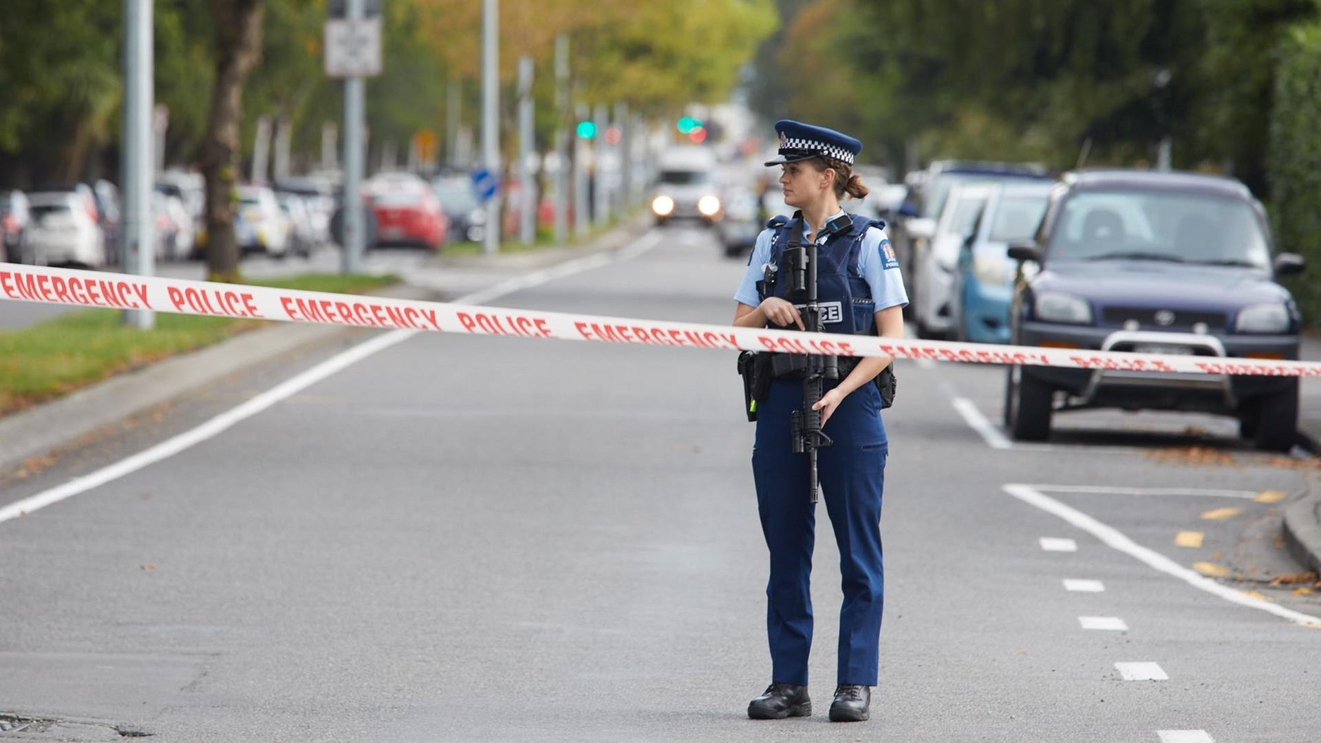 Eine Polizistin trägt ein Gewehr und steht hinter einer Polizeiabsperrung auf einer Straße. Eine Polizistin trägt ein Gewehr und steht hinter einer Polizeiabsperrung auf einer Straße.