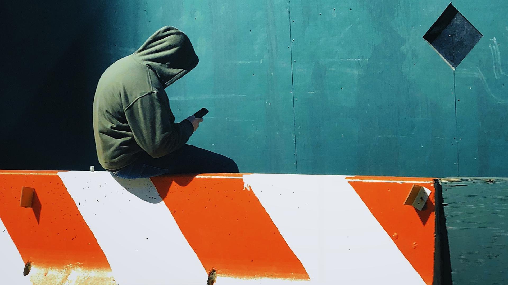 Ein junger Mensch mit Kapuzenshirt und Smartphone in der Hand sitzt auf einer Barriere.