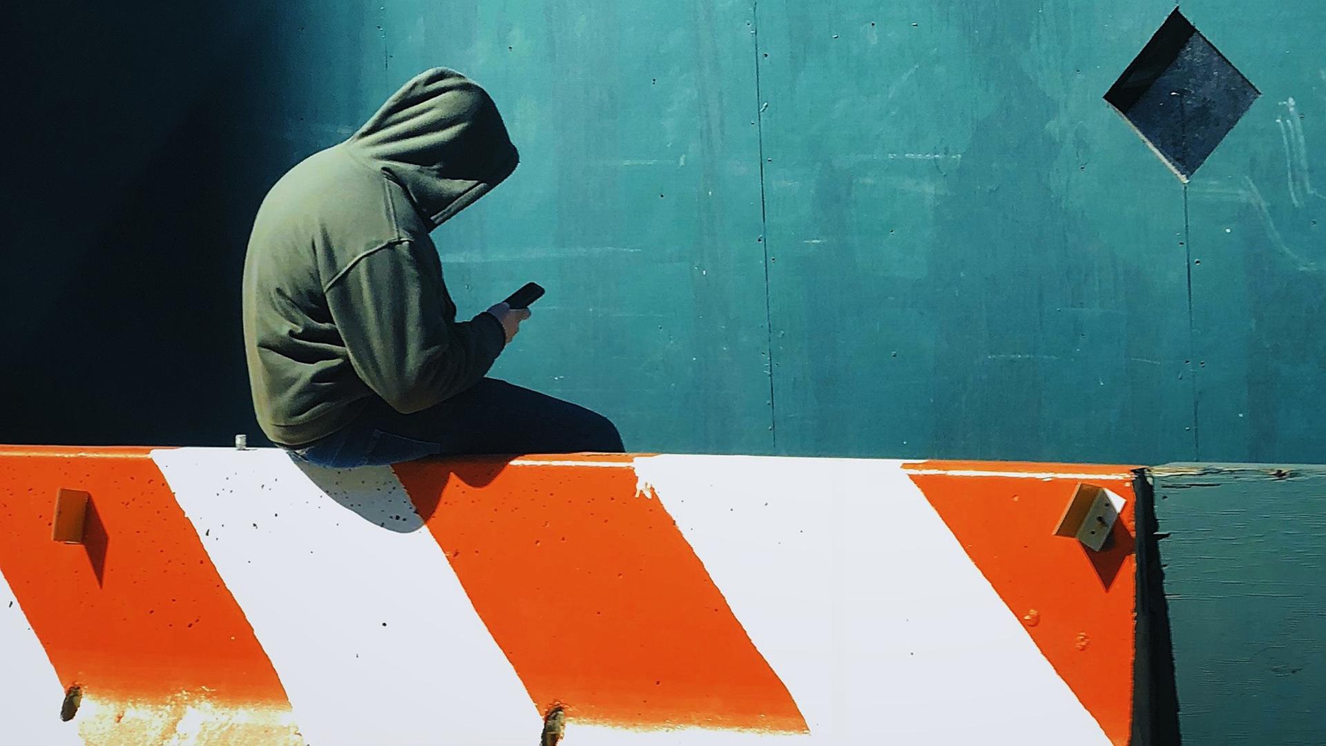 Ein junger Mensch mit Kapuzenshirt und Smartphone in der Hand sitzt auf einer Barriere. Ein junger Mensch mit Kapuzenshirt und Smartphone in der Hand sitzt auf einer Barriere.