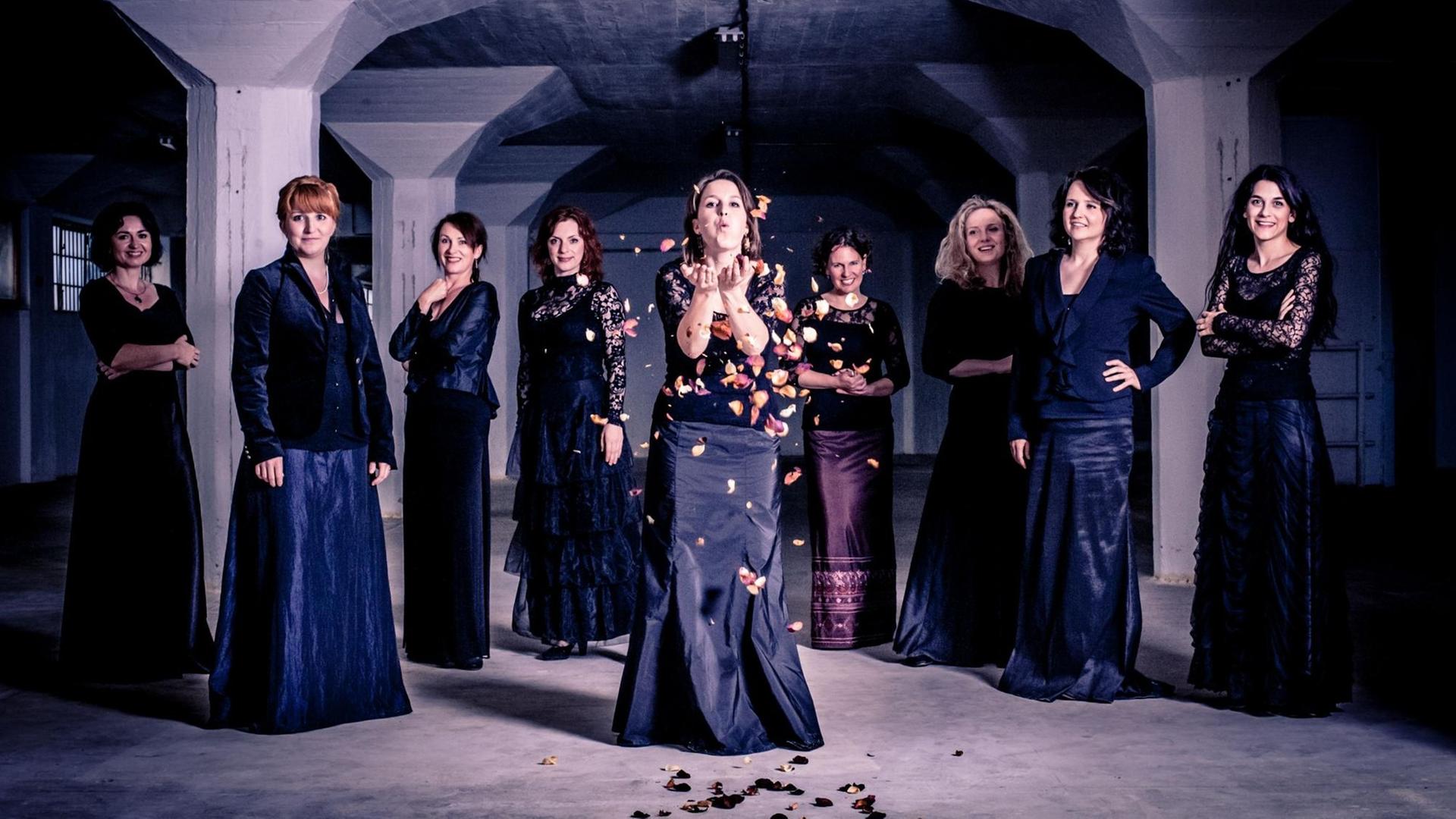Neun Sängerinnen stehen in einem Halbkreis in einem Keller-Gewölbe und tragen schwarze Kleidung. Eine von ihnen, vorne in der Mitte, pustet bunte Blätter in die Luft. 