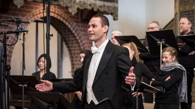 Der Kantor singt in einer Kirche gemeinsam mit einem Chor, der hinter ihm steht. Der Kantor singt in einer Kirche gemeinsam mit einem Chor, der hinter ihm steht.