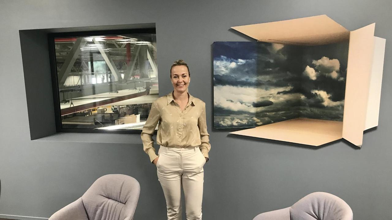 Eine junge Frau, die Hände in den Hosentaschen, steht zwischen zwei Bürostühlen vor einem Fenster mit Blick in den Betrieb. Eine junge Frau, die Hände in den Hosentaschen, steht zwischen zwei Bürostühlen vor einem Fenster mit Blick in den Betrieb.