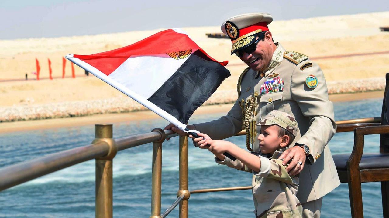 Präsident Sisis auf einem Schiff auf dem Suez-Kanal. Neben ihm steht ein Kind - ebenfalls in Uniform - und schwenkt die ägyptische Flagge. 