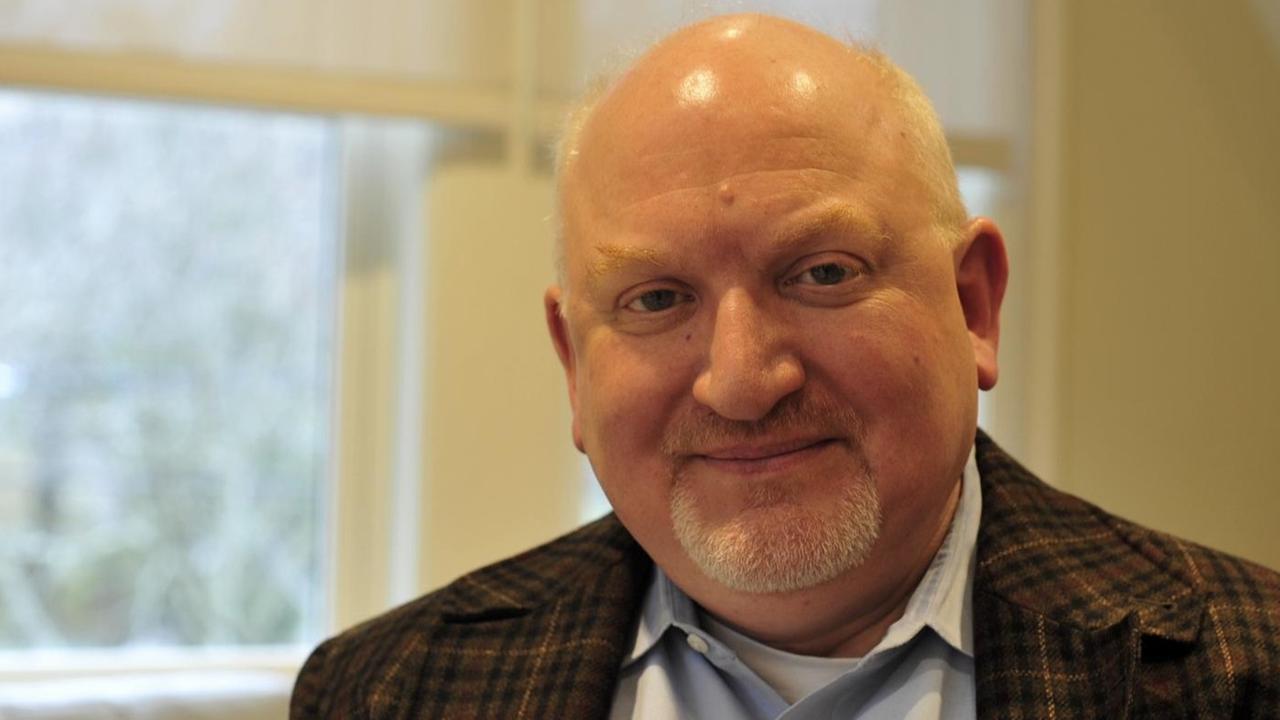 Professor Martin Jean, im Büro, mit kariertem Sakko in offenem weißem Hemd, lächelt freundlich in die Kamera. Professor Martin Jean, im Büro, mit kariertem Sakko in offenem weißem Hemd, lächelt freundlich in die Kamera.