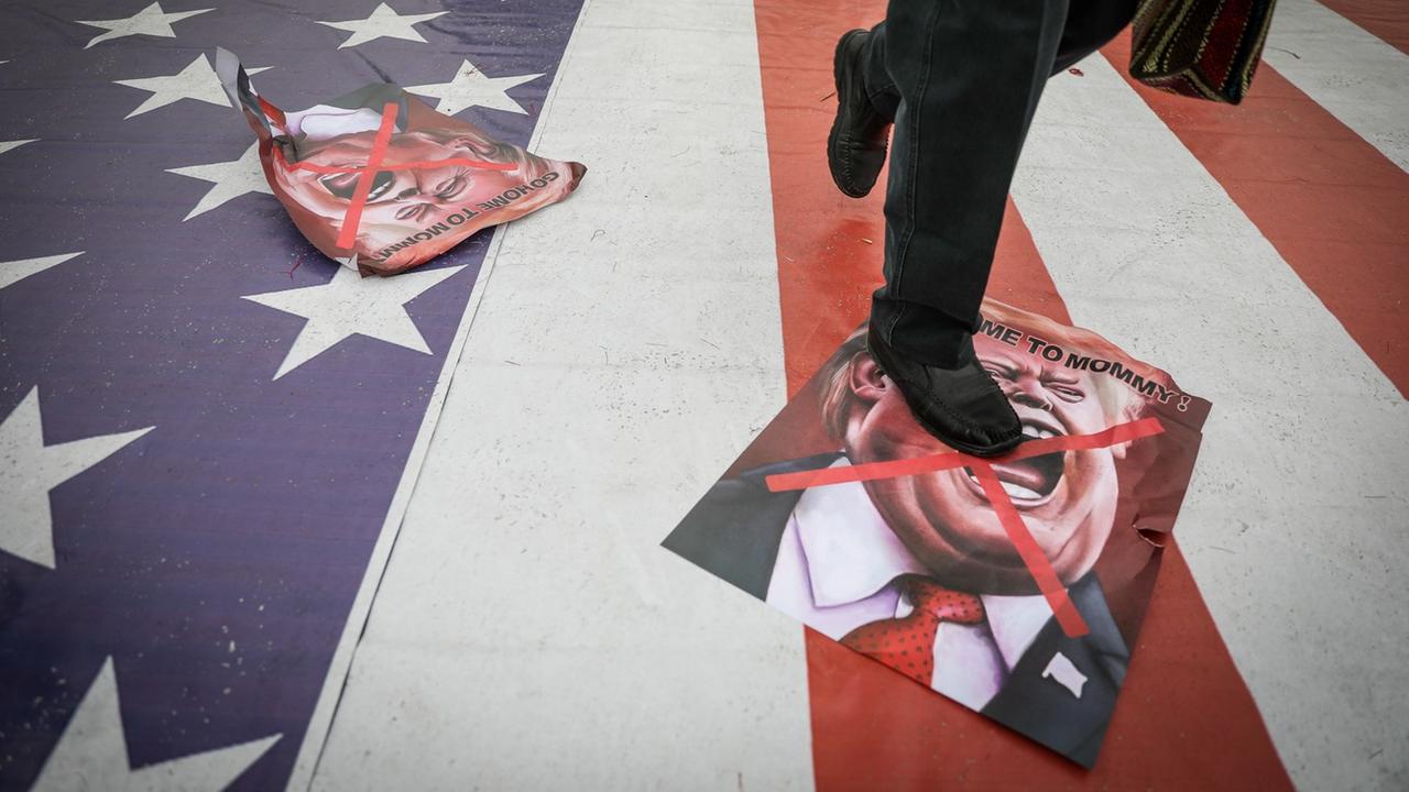 Plakate mit einer Donald Trump Karikatur liegen auf dem Boden eine Bürgersteigs in Teheran, der mit der amerikanischen Flagge bemalt ist. Darauf läuft eine Person, wobei das Bild am Rumpf endet. Plakate mit einer Donald Trump Karikatur liegen auf dem Boden eine Bürgersteigs in Teheran, der mit der amerikanischen Flagge bemalt ist. Darauf läuft eine Person, wobei das Bild am Rumpf endet.