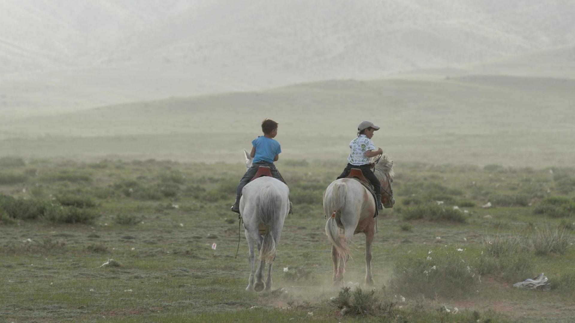 Zwei mongolische Kinder reiten im trockenen Grasland auf weißen mongolischen Pferden.