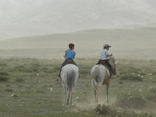 Zwei mongolische Kinder reiten im trockenen Grasland auf weißen mongolischen Pferden.