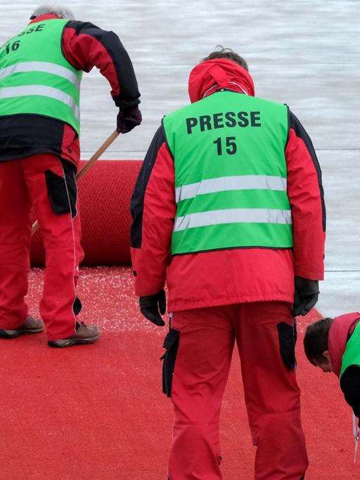Drei Arbeiter in grünen Warnwesten mit der Aufschrift "Presse" arbeiten an dem fast komplett aufgerollten Teppich. Einer fegt Schneegraupel weg.