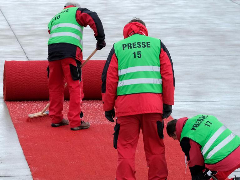 Drei Arbeiter in grünen Warnwesten mit der Aufschrift "Presse" arbeiten an dem fast komplett aufgerollten Teppich. Einer fegt Schneegraupel weg.