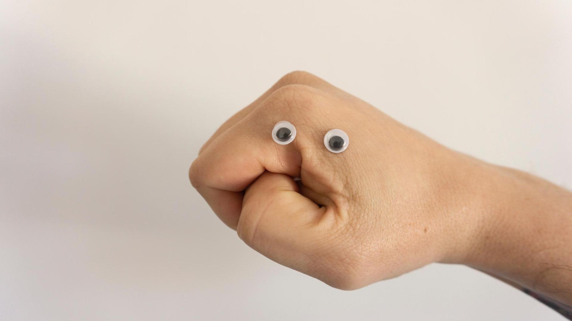 Eine Hand ist zu einer Faust geformt. Zwei Wackelaugen aus Plastik liegen zwischen Zeigefinger und Daumen. Eine Hand ist zu einer Faust geformt. Zwei Wackelaugen aus Plastik liegen zwischen Zeigefinger und Daumen.