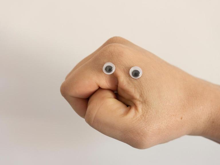 Eine Hand ist zu einer Faust geformt. Zwei Wackelaugen aus Plastik liegen zwischen Zeigefinger und Daumen.