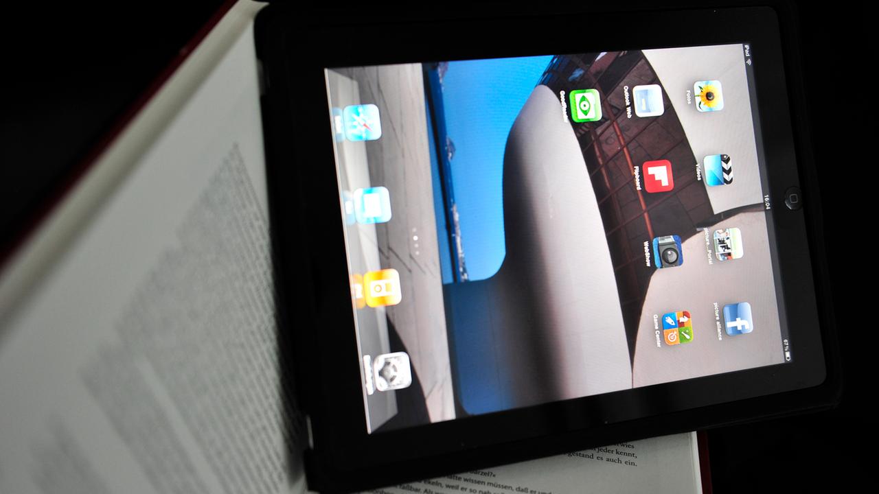 Ein iPad mit Apps auf dem Bildschirm liegt auf einem aufgeschlagenen Buch. Ein iPad mit Apps auf dem Bildschirm liegt auf einem aufgeschlagenen Buch.