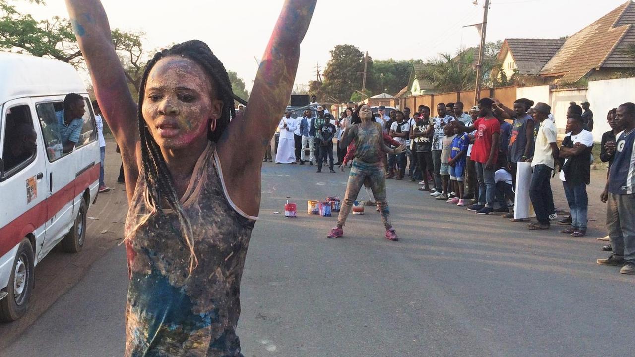 Straßenperformance der kongolesischen Künstlerin Sarah Mukadi. Sie ist am ganzen Körper bunt bemalt und hebt die Arme. Straßenperformance der kongolesischen Künstlerin Sarah Mukadi. Sie ist am ganzen Körper bunt bemalt und hebt die Arme.