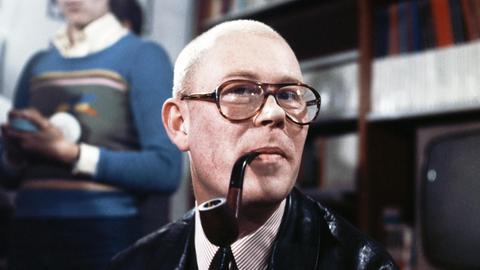 Schriftsteller Uwe Johnson mit Hornbrille und einer Pfeife im Mund bei einer Lesung, ca. Mitte der 70er-Jahre.