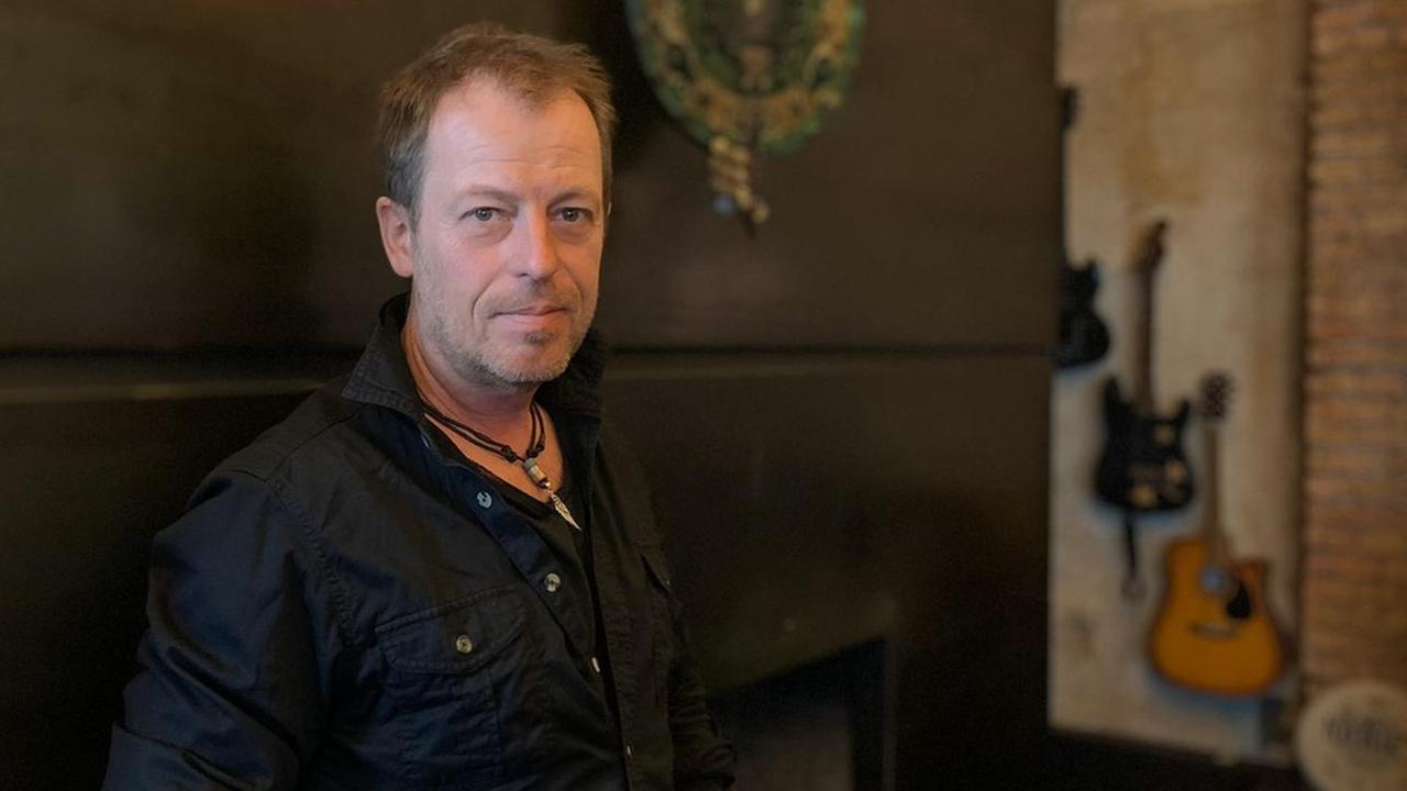 Porträt von Sascha Disselkamp: Er steht in schwarzer Kleidung vor einem dunklen Hintergrund, etwas abseits hängen drei Gitarren an der Wand. Porträt von Sascha Disselkamp: Er steht in schwarzer Kleidung vor einem dunklen Hintergrund, etwas abseits hängen drei Gitarren an der Wand.