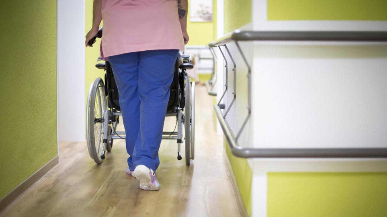 Eine Altenpflegerin schiebt eine Bewohnerin in einem Altenpflegeheim in einem Rollstuhl den Gang entlang. 