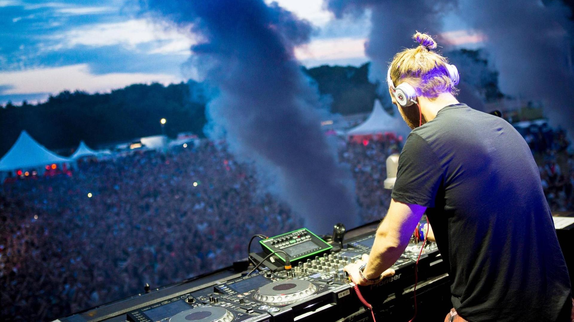 David Guetta von hinten bei einem Konzert auf der Hamburger Trabrennbahn. Vor ihm ein riesiges Mischpult, dahinter Dampf-Fontänen eines Special Effects und dann tausende Menschen im Publikum David Guetta von hinten bei einem Konzert auf der Hamburger Trabrennbahn. Vor ihm ein riesiges Mischpult, dahinter Dampf-Fontänen eines Special Effects und dann tausende Menschen im Publikum