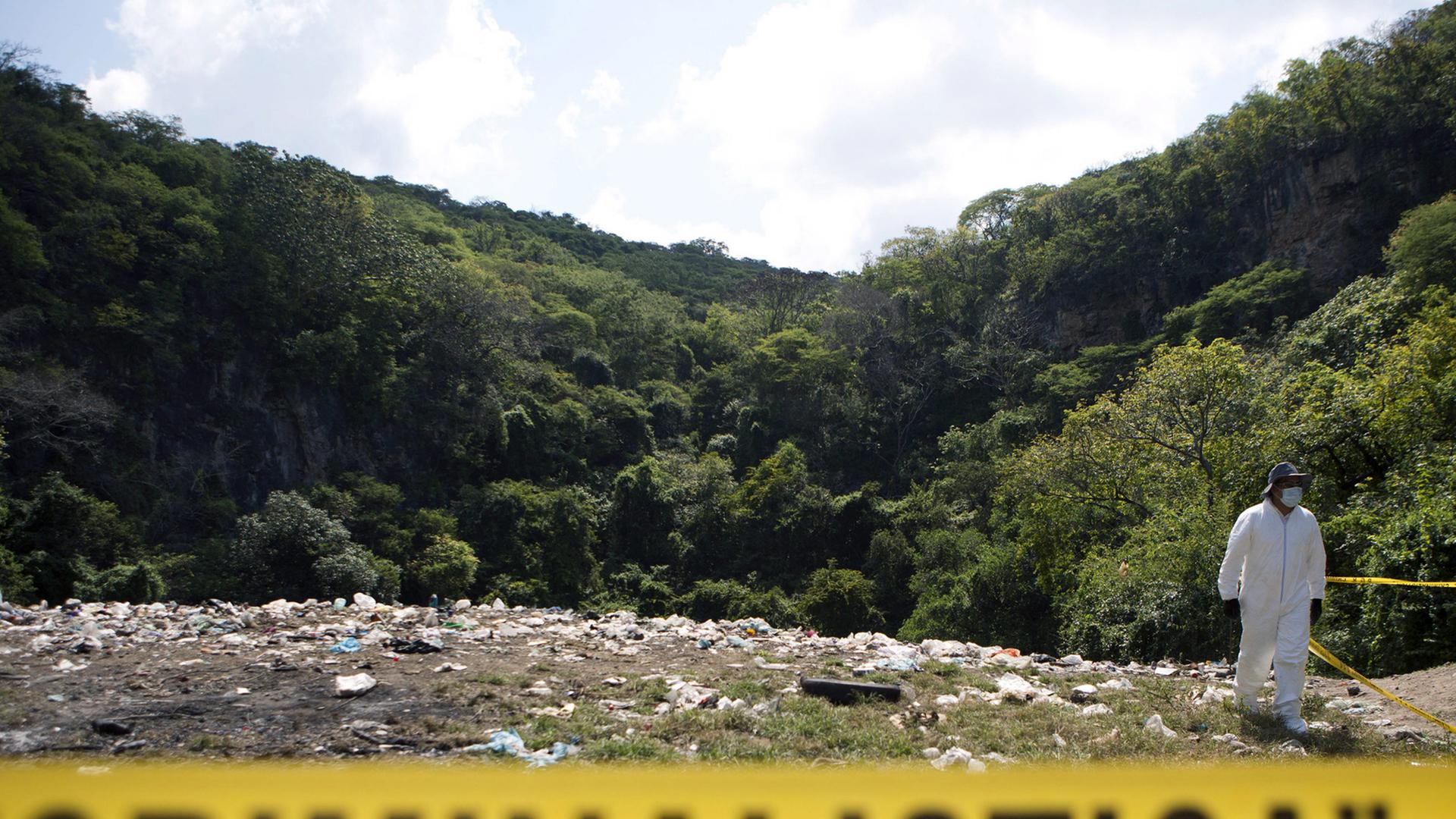 Hinter einer gelben Absperrung mit der schwarzen Aufschrift "Criminalistica" sieht man eine Müllkippe mit einem in weiße Schutzkleidung gehüllten Forensiker, im Hintergrund ein bewaldeter Hügel.