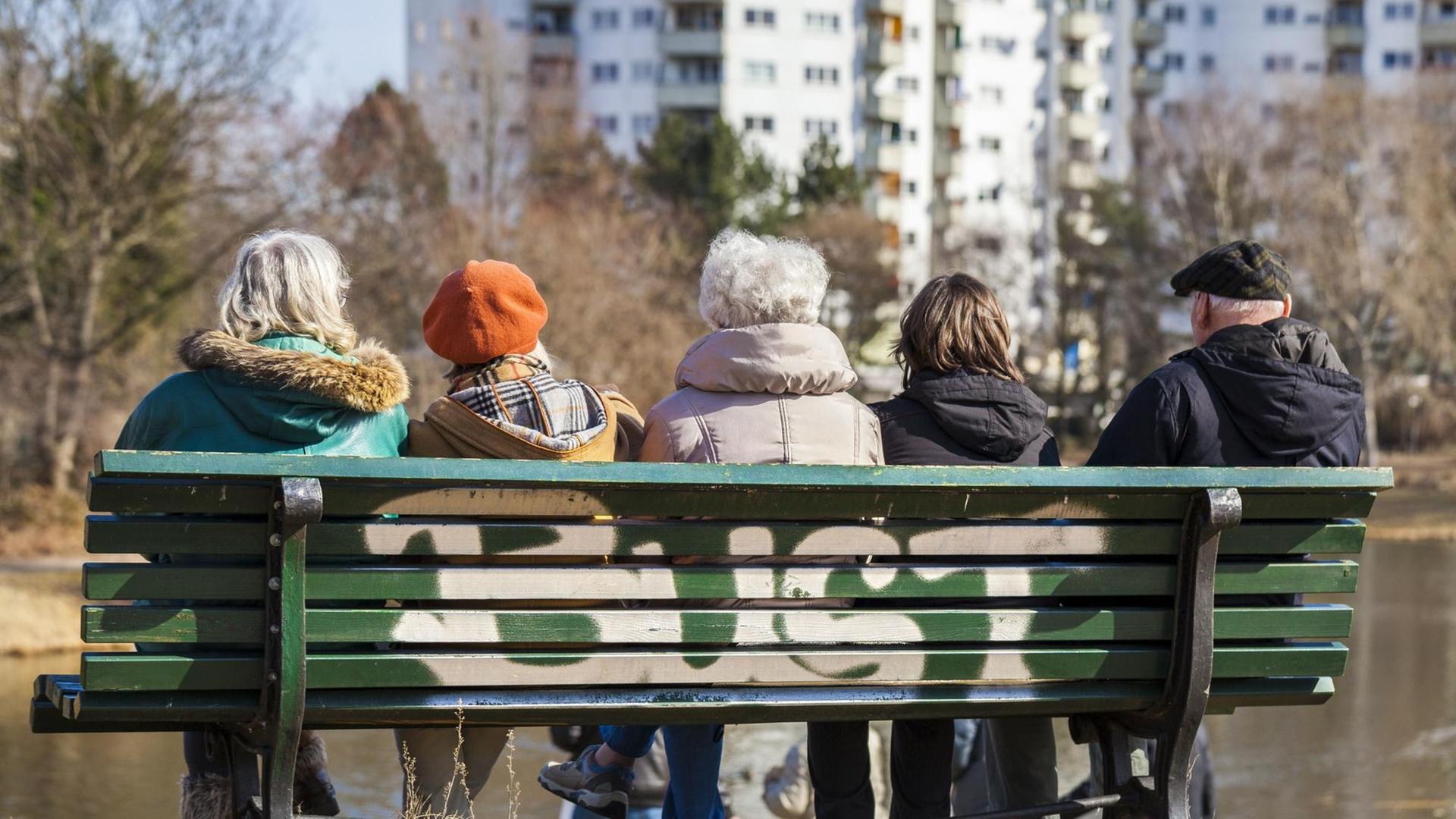 Mehrere Senioren sitzen auf einer Bank, im Hintergrund ist ein großer Wohnungsblock zu sehen.