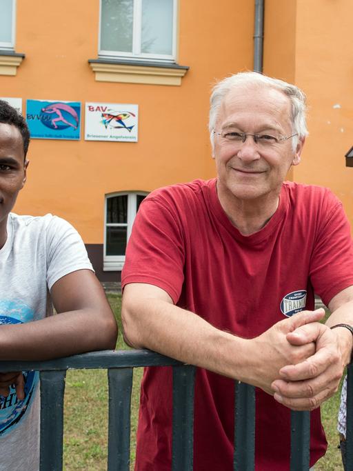 Der Bundestagsabgeordnete Martin Patzelt (CDU) und die beiden Flüchtlinge aus Eritrea Haben und Awet am Zaun des Gemeinde- und Vereinshauses in Briesen im Landkreis Oder-Spree (Brandenburg). Sie lehnen sich über ein Geländer und schauen in die Kamera.