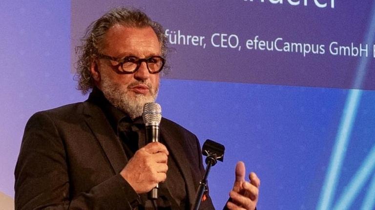Thomas Anderer, Geschäftsführer von efeuCampus, hält einen Vortrag. Auf der Leinwand hinter ihm ist eine Präsentation zu sehen. Thomas Anderer, Geschäftsführer von efeuCampus, hält einen Vortrag. Auf der Leinwand hinter ihm ist eine Präsentation zu sehen.