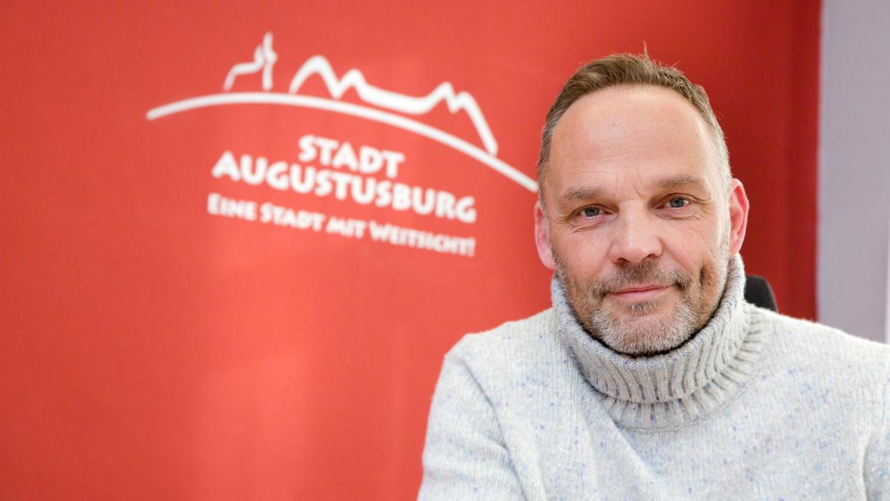 Dirk Neubauer vor einem roten Hintergrund, auf dem Stadt Augustusburg steht. Er trägt einen hellen Rollkragenpullover und hat einen Vollbart.