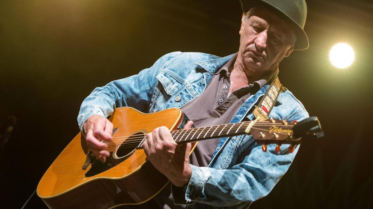 Ringsgwandl steht in Jeansjacke auf einer Bühne und spielt Gitarre. Ringsgwandl steht in Jeansjacke auf einer Bühne und spielt Gitarre.