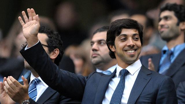 Scheich Mansour bin Zayed Al Nahyan steht im Anzug winkend und lächelnd auf einer Tribüne, im Hintergrund sind weitere Männer zu sehen