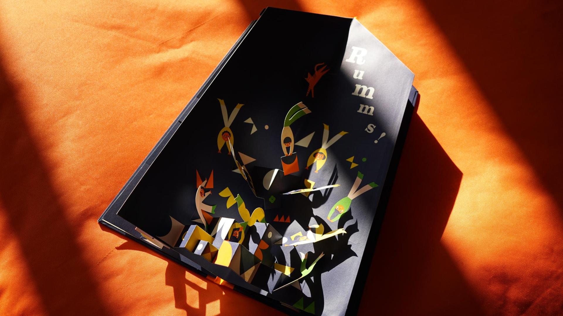 Das aufgeschlagene Kinderbuch "Eins, zwei, drei, die Akrobaten" auf orangenem Hintergrund.
