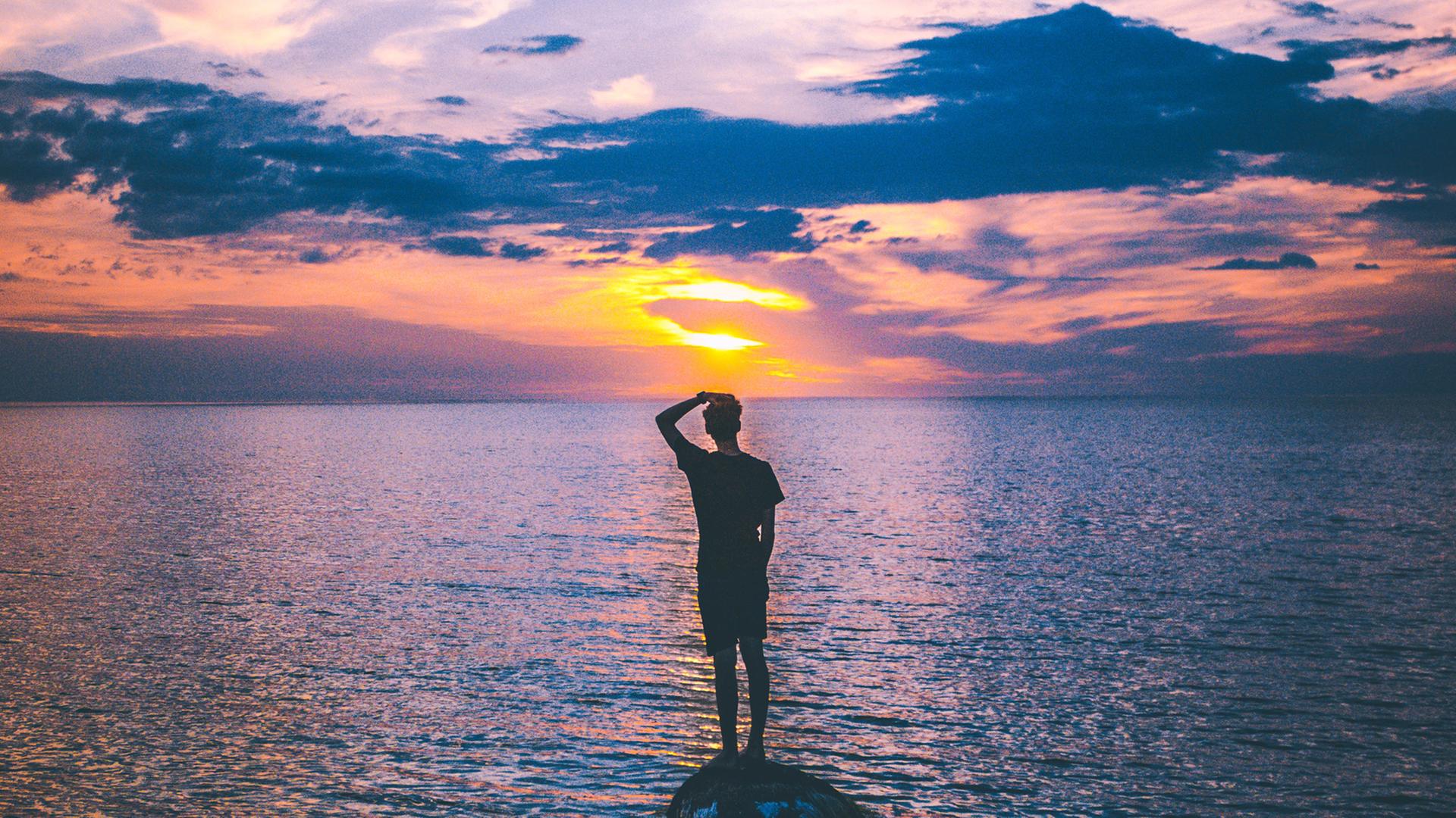 Ein Mann steht auf einem kleinen Felsen im Meer und schaut in die Abendsonne.
