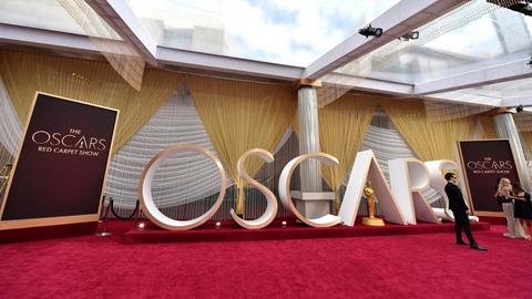 Blick auf den "Oscars"-Schriftzug, der 2020 in einem Raum auf einem roten Teppich stand. Daneben Schilder mit der Aufschrift "The Oscars Red Carpet Show", eine Vorabshow vor der Verleihung der Oscars. Am Rand stehen in Abendrobe gekleidete Personen.