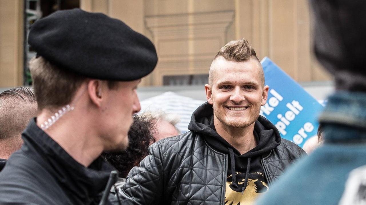 Der rechtsextrem Rapper Chris Ares in einem schwarzen Sweatshirt mit goldenem Adler drauf in einer Menschenmenge mit Transparenten. Im Vordergrund ein Einsatzpolizist in Uniform. Der rechtsextrem Rapper Chris Ares in einem schwarzen Sweatshirt mit goldenem Adler drauf in einer Menschenmenge mit Transparenten. Im Vordergrund ein Einsatzpolizist in Uniform.