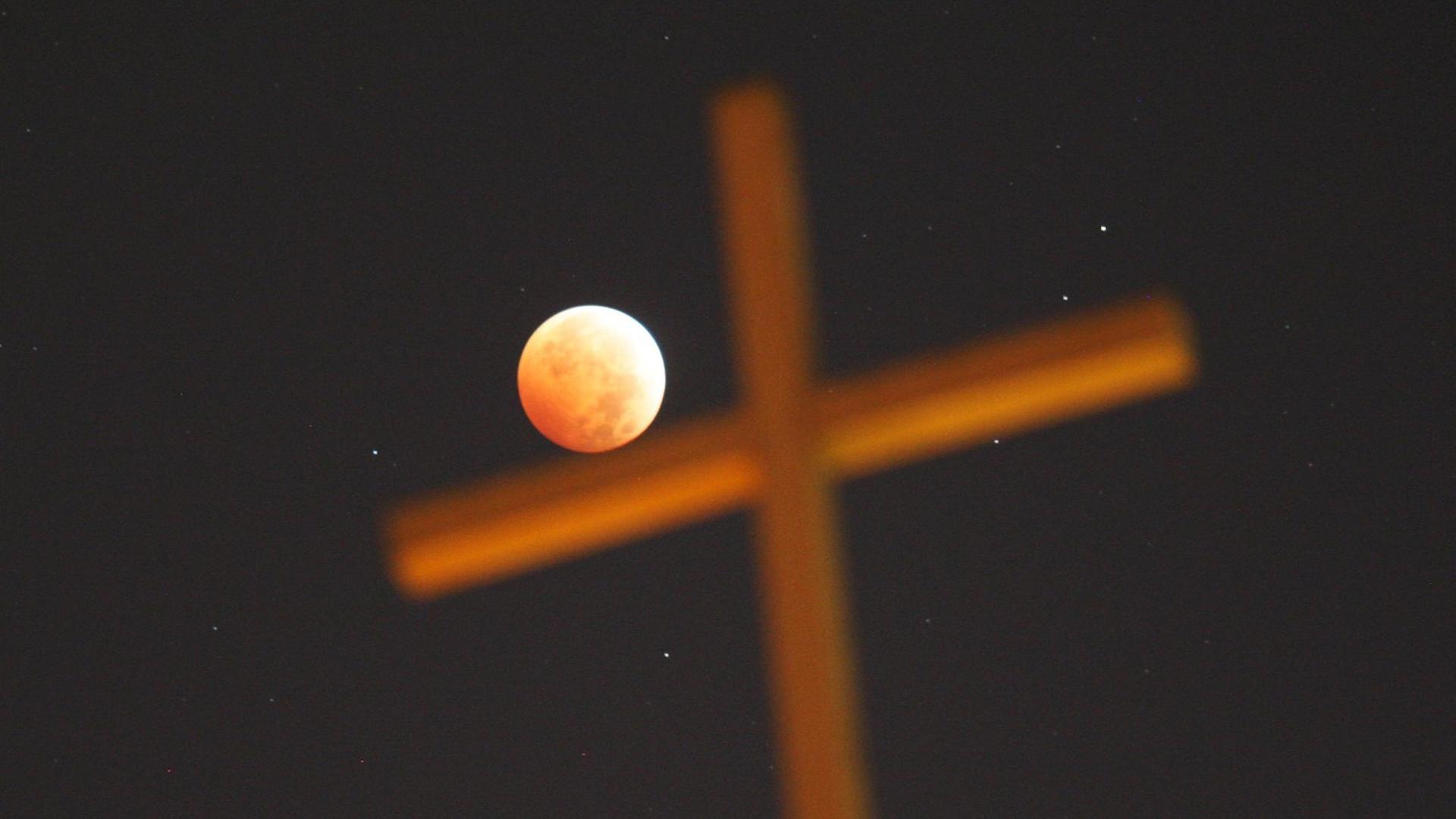 Ein religiöses Kreuz vor dem Nachhimmel, während einer totalen Mondfinsternis. Der rot erleuchtete Mond scheint das Kreuz zu berühren. Ein religiöses Kreuz vor dem Nachhimmel, während einer totalen Mondfinsternis. Der rot erleuchtete Mond scheint das Kreuz zu berühren.