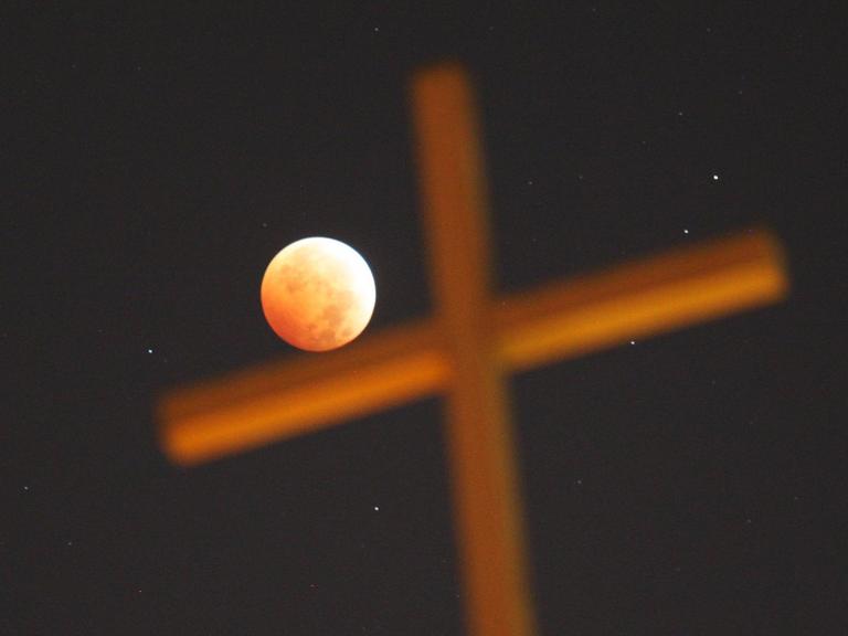 Ein religiöses Kreuz vor dem Nachhimmel, während einer totalen Mondfinsternis. Der rot erleuchtete Mond scheint das Kreuz zu berühren.