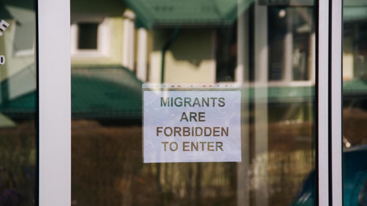 An einer Glastür zu einem Supermarkt ist ein Schild angebracht, auf dem steht "Migrants are forbidden to enter" - Migranten dürfen hier nicht hinein. An einer Glastür zu einem Supermarkt ist ein Schild angebracht, auf dem steht "Migrants are forbidden to enter" - Migranten dürfen hier nicht hinein.