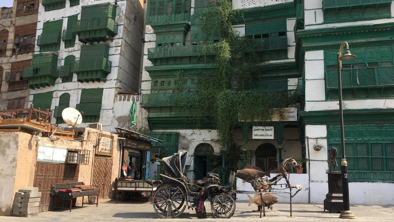 In der Altstadt von Jeddah, vor Häusern mit den typischen grünen Holzvorbauten vor den Fenstern, steht eine Skulptur aus rostigem Metall in Gestalt einer Kutsche mit Pferd, flankiert von zwei großen Fischen. In der Altstadt von Jeddah, vor Häusern mit den typischen grünen Holzvorbauten vor den Fenstern, steht eine Skulptur aus rostigem Metall in Gestalt einer Kutsche mit Pferd, flankiert von zwei großen Fischen.
