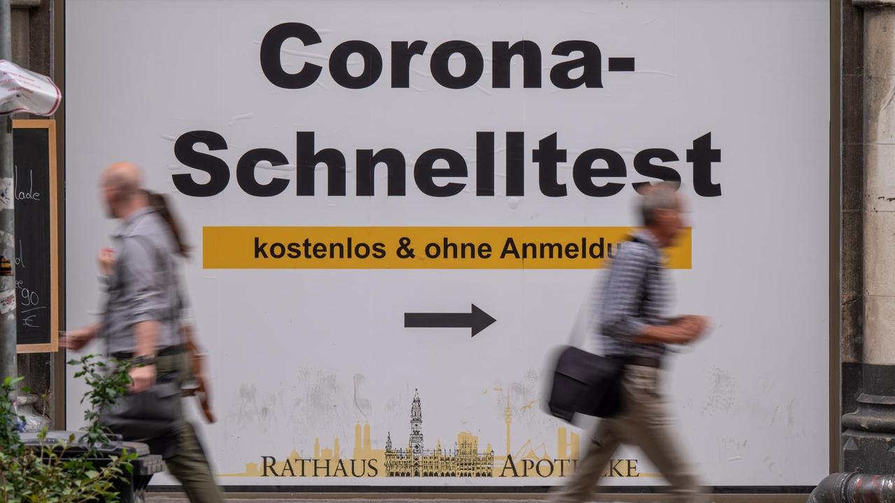 Passanten gehen in der Fußgängerzone in der Innenstadt an einem Plakat mit der Aufschrift „Corona-Schnelltest - kostenlos und ohne Anmeldung“ vorbei.
