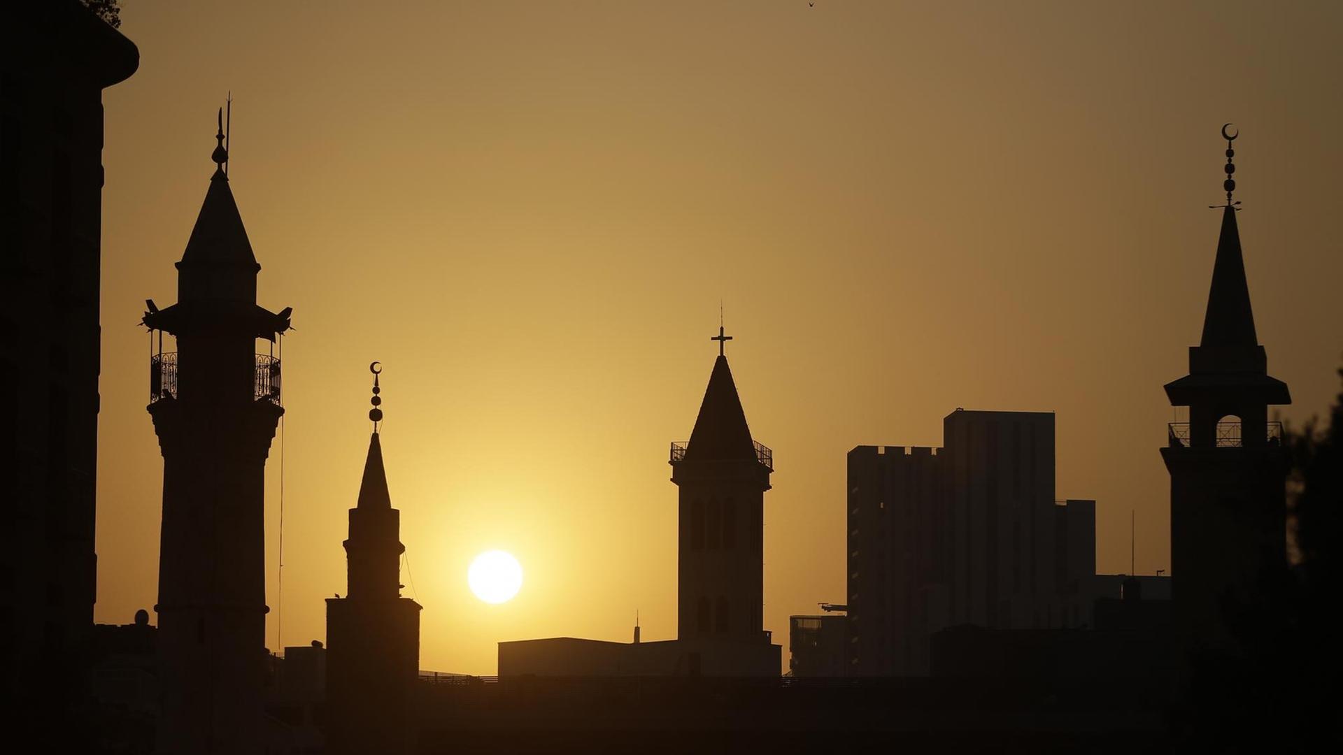 Die Dächer von Kirchen und Moscheen heben sich als Silhouetten vor einem Sonnenuntergang ab.