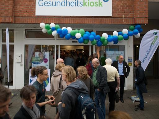 Passanten und Teilnehmer der Eröffnungsfeier stehen am 28.09.2017 vor dem neuen Gesundheitskiosk in Hamburg. In zentraler Lage wird das mehrsprachige Kiosk-Team aus Gesundheitsfachkräften die Bürger aus den Stadtteilen Billstedt und Horn kostenlos zu allen Fragen der Gesundheit beraten. Foto: Daniel Reinhardt/dpa | Verwendung weltweit
