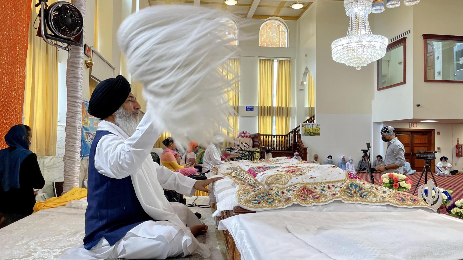 In der Gebetshalle eines New Yorker Sikh-Tempels wird das heilige Buch Guru Grant Sahib unter einem roten Baldachin präsentiert. Es ist mit einem bestickten Tuch zugedeckt, dahinter sitzt ein Sikh mit schwarzem Turban und blauer Weste und fächelt dem Buch mit einer Art weißem Federbusch Luft zu.