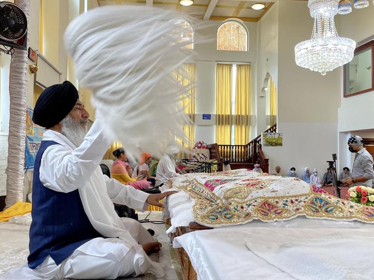 In der Gebetshalle eines New Yorker Sikh-Tempels wird das heilige Buch Guru Grant Sahib unter einem roten Baldachin präsentiert. Es ist mit einem bestickten Tuch zugedeckt, dahinter sitzt ein Sikh mit schwarzem Turban und blauer Weste und fächelt dem Buch mit einer Art weißem Federbusch Luft zu.