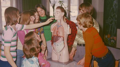 Biologieunterricht in einer Schule in Neubrandenburg (DDR): Die menschliche Anatomie wird anhand eines Modells erlernt. Die Schülerinnen und Schüler berühren vorsichtig das Modell eines Menschen und blicken neugierig auf das Gesicht, 1975.