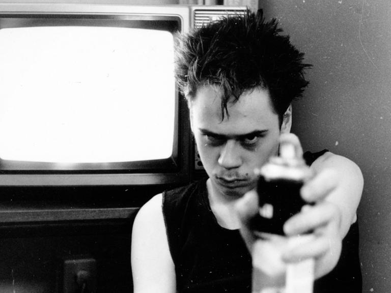 Schwarz-Weiß Aufnahme eines Mannes in schwarzem Tanktop vor einem Fernseher mit einer Sprühflasche in der Hand.
