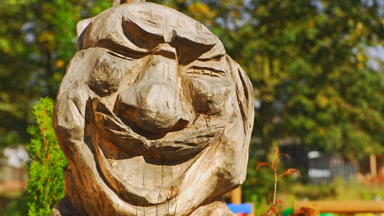 Der Kopf eines freundlichen Dämons, mit groben Schnitten aus einem Baumstamm geschnitzt, zeigt ein rundes, lächelndes Gesicht mit Schnauzbart und Halbglatze. Aufnahme im Freien, auf einem sonnenbeschienenen Platz mit Bäumen im Hintergrund.