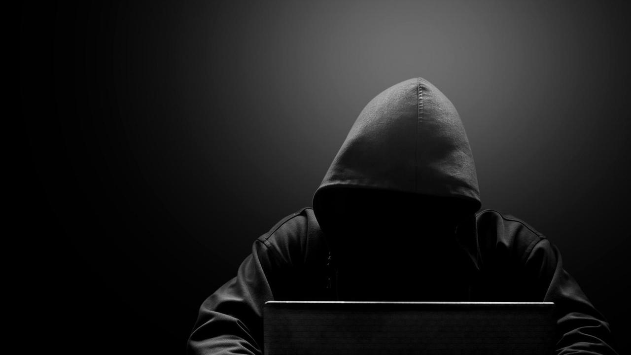 Ein Mann mit schwarzer Kapuze und verschattetem Gesicht sitzt vor einem Laptop. Ein Mann mit schwarzer Kapuze und verschattetem Gesicht sitzt vor einem Laptop.