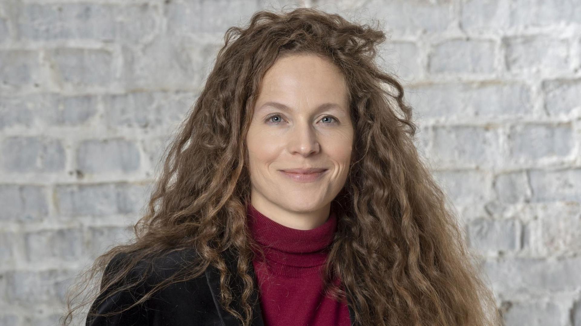 Portrait einer Frau mit langen dunkelblonden Locken, die vor einer weißen Mauer steht und direkt in die Kamera blickt. Portrait einer Frau mit langen dunkelblonden Locken, die vor einer weißen Mauer steht und direkt in die Kamera blickt.