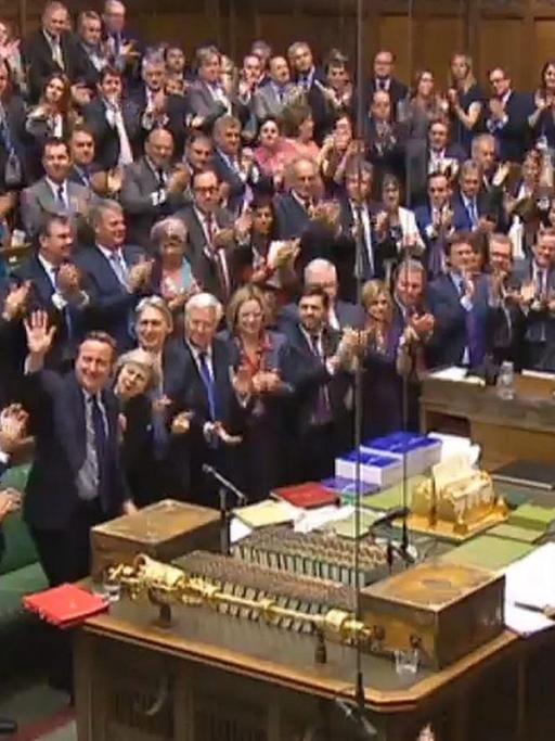 Das Bild zeigt das vollbesetze applaudierende Unterhaus; Cameron steht am Rednerpult und winkt den Abgeordneten zu.