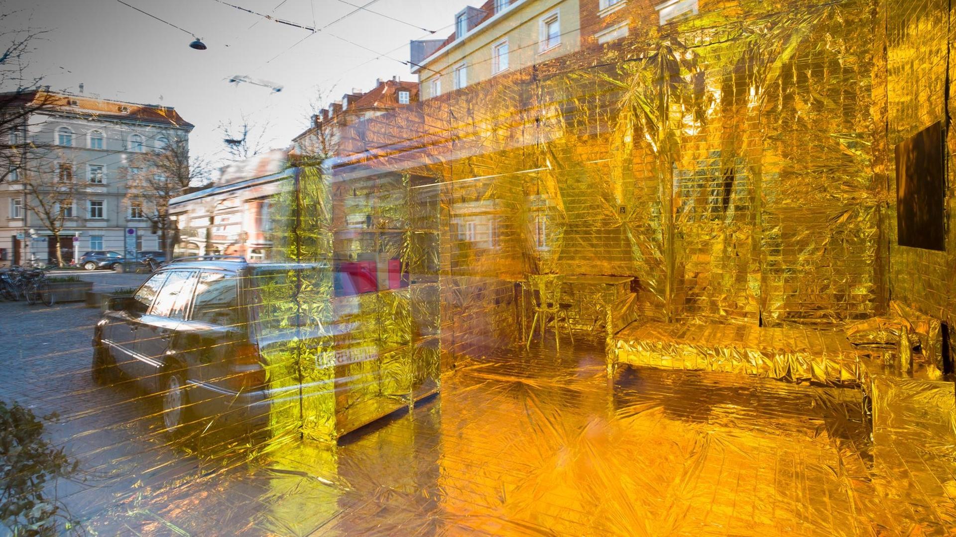 Ein in goldschimmernde Folie verpacktes Zimmer samt Einrichtung ist am 26.03.2017 in München (Bayern) im Stadtteil Haidhausen durch die Scheibe eines umgebauten Ladengeschäfts zu sehen. In der Schaufensterscheibe spiegelt sich der Genoveva-Schauer-Platz. Die Installation gehört zu einer Kunstaktion, die auf den immer teurer werdenden Wohnraum in der bayerischen Landeshauptstadt aufmerksam machen soll. (zu dpa "Vergoldetes Zimmer als Protest gegen Wohnungsnot" vom 28.03.2017) Foto: Peter Kneffel/dpa | Verwendung weltweit Ein in goldschimmernde Folie verpacktes Zimmer samt Einrichtung ist am 26.03.2017 in München (Bayern) im Stadtteil Haidhausen durch die Scheibe eines umgebauten Ladengeschäfts zu sehen. In der Schaufensterscheibe spiegelt sich der Genoveva-Schauer-Platz. Die Installation gehört zu einer Kunstaktion, die auf den immer teurer werdenden Wohnraum in der bayerischen Landeshauptstadt aufmerksam machen soll. (zu dpa "Vergoldetes Zimmer als Protest gegen Wohnungsnot" vom 28.03.2017) Foto: Peter Kneffel/dpa | Verwendung weltweit