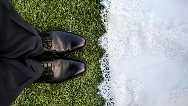 Links schwarze Hose, schwarze Schuhe, rechts der untere Teil eines weißen Kleides. Darunter grüne Wiese.